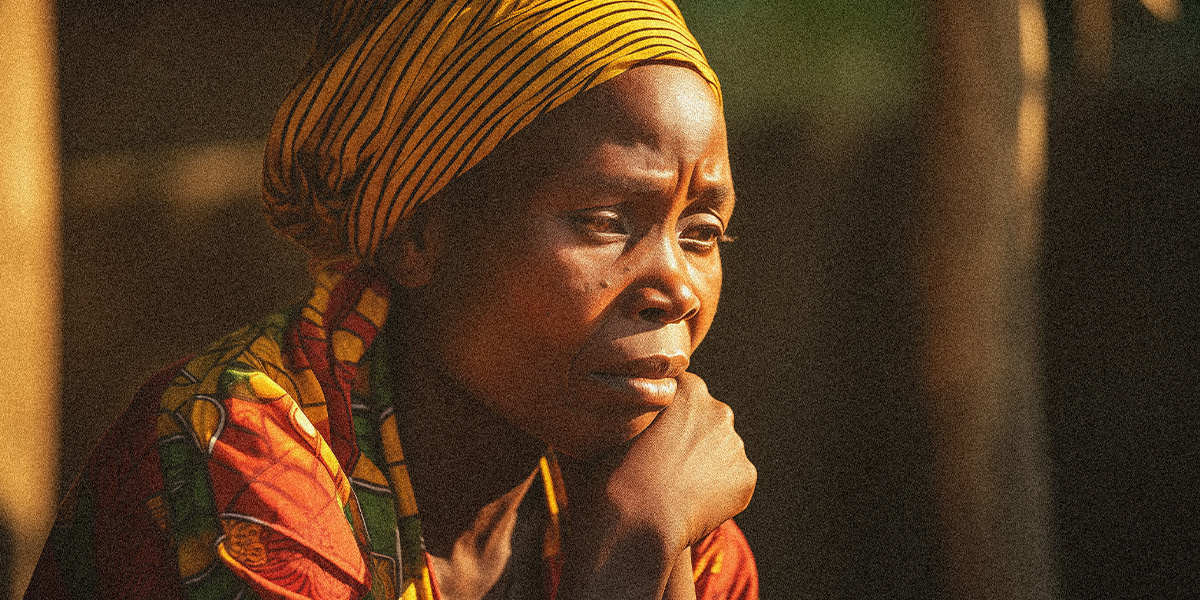 An African woman sitting looking as if she's deep in thought wearing a black stripped and yellow headress and a warmly patterened and bright shirt.