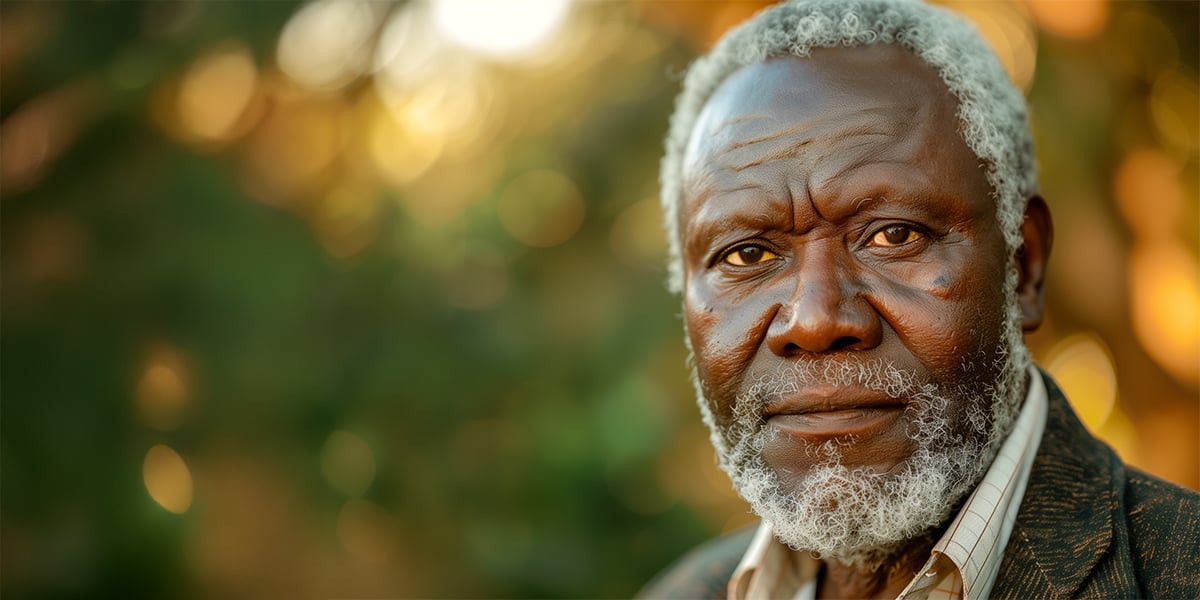 Older African gentleman wearing a corduroy sport coat and a collared shirt . He's standing outdoors in a shaded area.